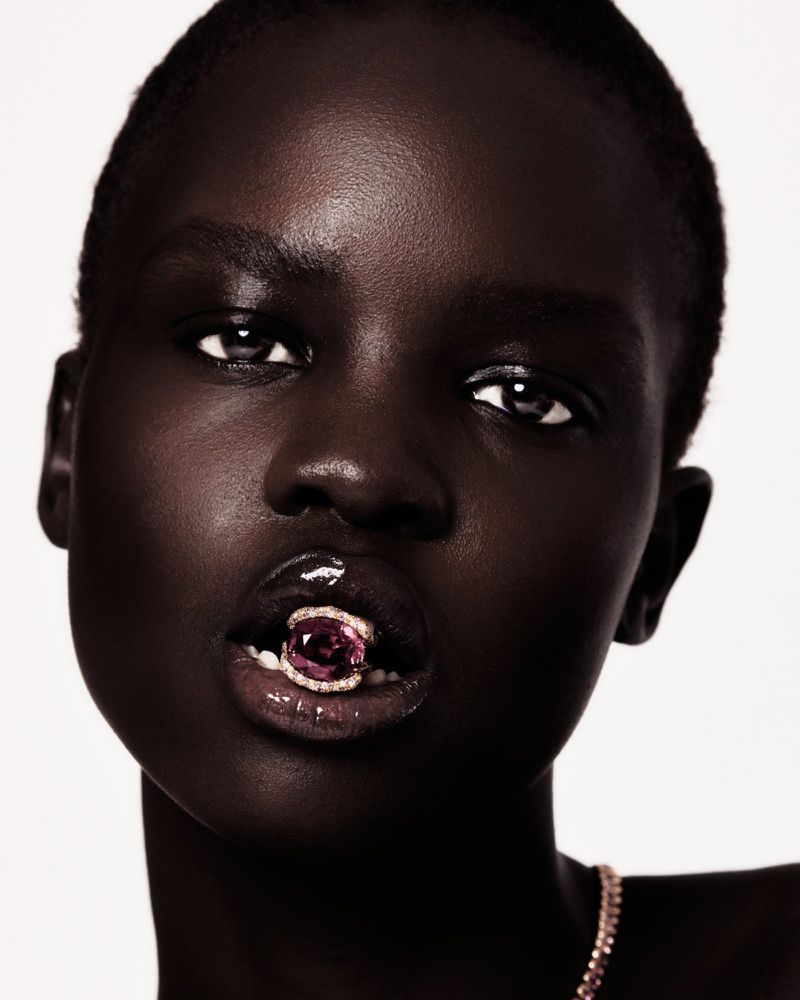 Close-up beauty shot of a dark-skinned adult female model with glossy lips, holding a jeweled ring in her mouth in a photo studio setting.