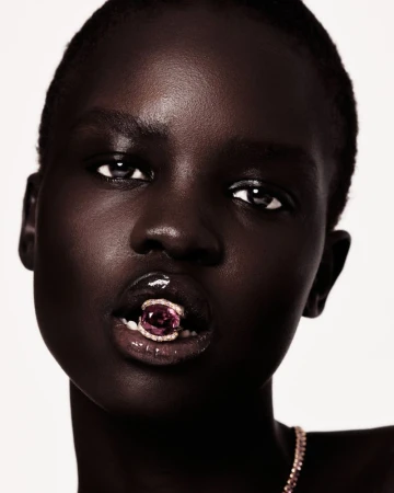 Close-up beauty shot of a dark-skinned adult female model with glossy lips, holding a jeweled ring in her mouth in a photo studio setting.