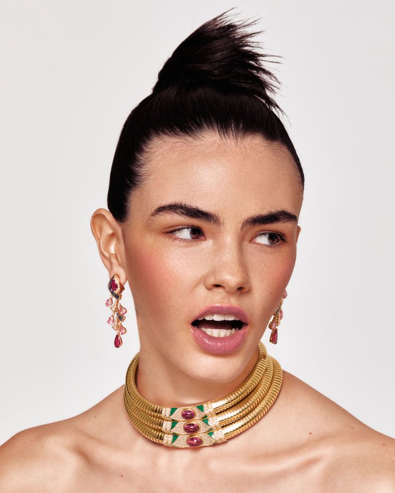 A striking beauty shot showcasing a woman with dark hair styled in a high bun, wearing gold jewelry and earrings against a bright backdrop.