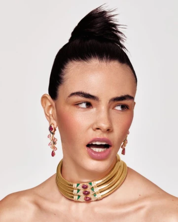 A striking beauty shot showcasing a woman with dark hair styled in a high bun, wearing gold jewelry and earrings against a bright backdrop.
