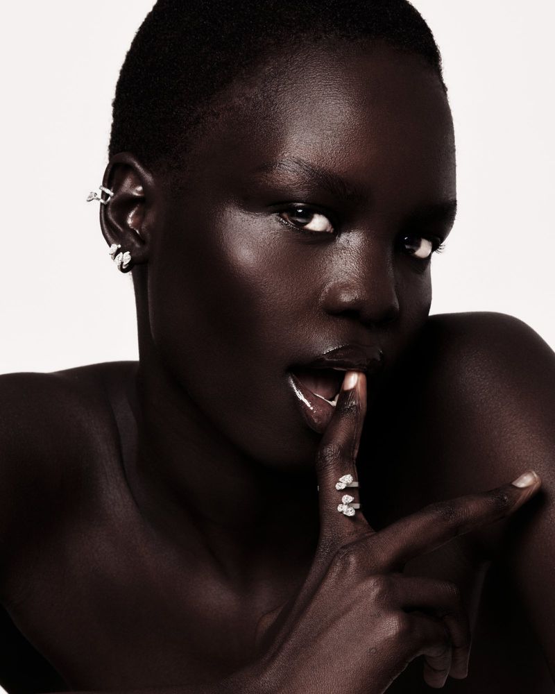 Close-up studio portrait of an adult woman with dark skin wearing ear cuffs and rings, her finger to her lips against a white background.
