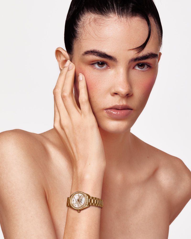Close-up of a young woman with slicked-back hair and flushed cheeks, wearing a gold wristwatch, posing in front of a white backdrop.