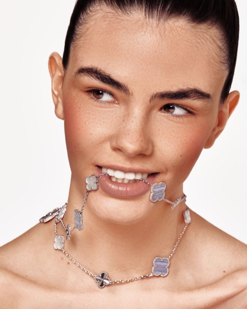 Smiling woman poses in a studio shot, wearing a silver necklace. The necklace has floral quatrefoil motifs, and the background is white.