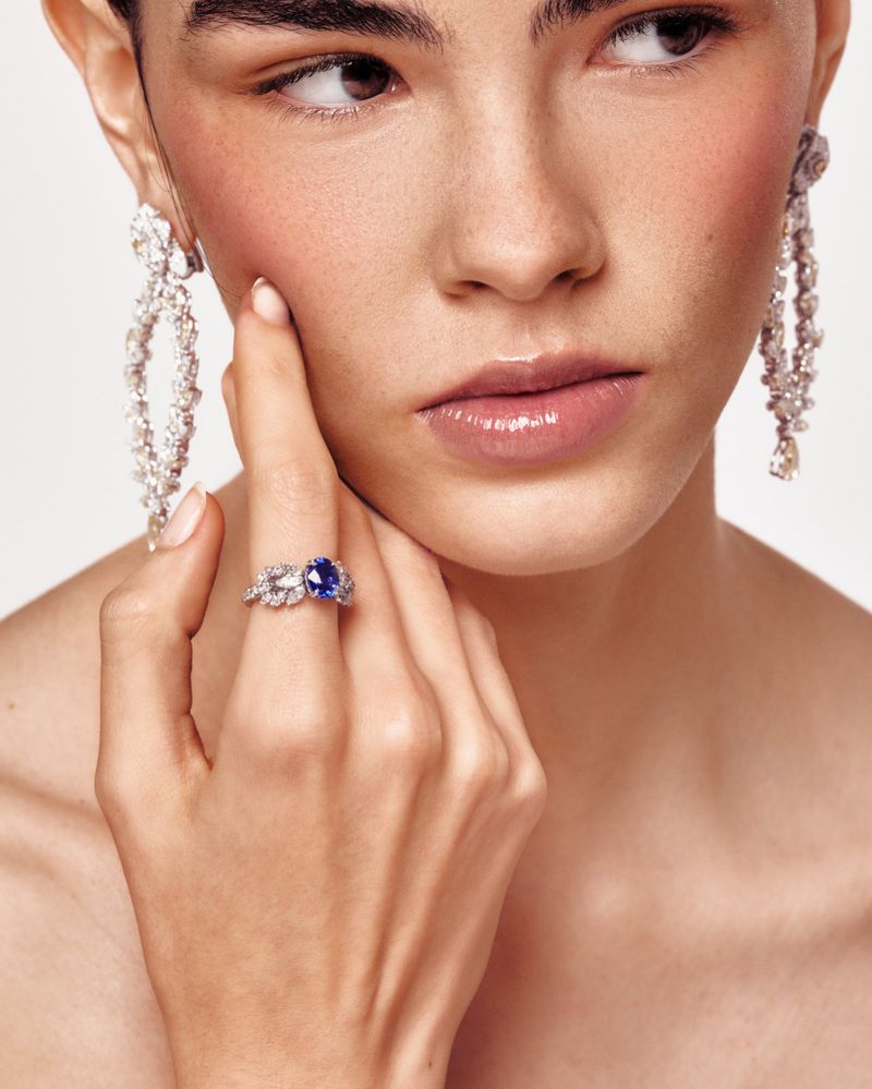 Close-up portrait of a woman wearing a ring, earrings, and makeup, gazing away from the camera in a softly lit, neutral-toned setting.