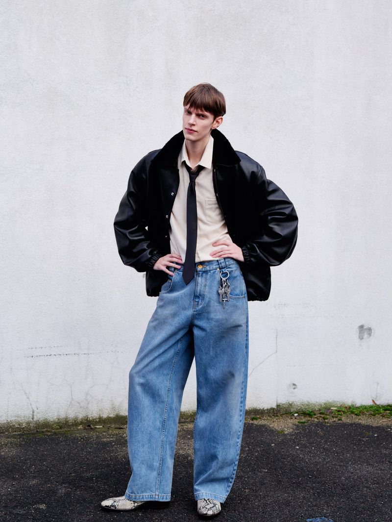 Model standing outdoors wearing an oversized black leather jacket, light shirt with tie, and loose blue jeans against a textured wall.