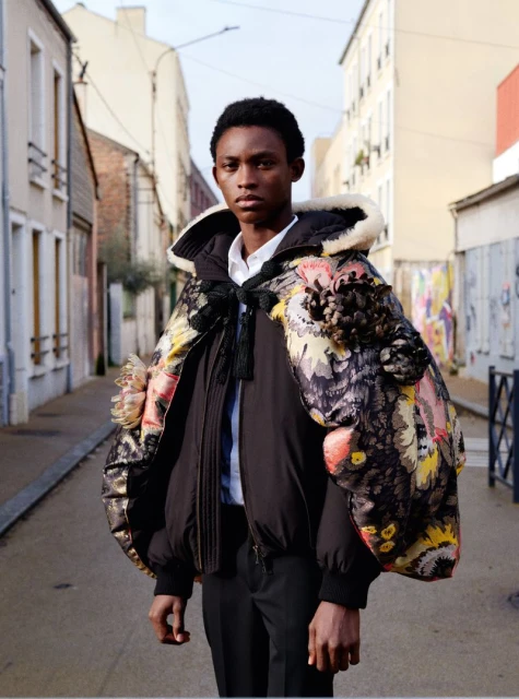 A young Black male model in a statement cape and black puffer jacket stands on an empty street, showcasing a unique fashion ensemble.
