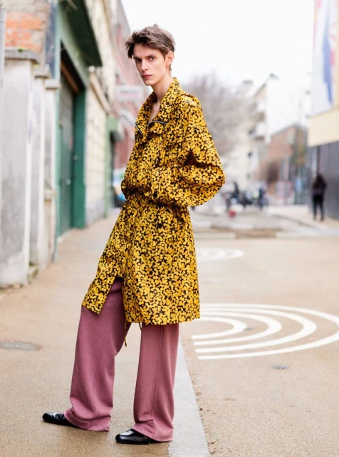Fashionable adult poses in a mustard-yellow and black floral coat with mauve trousers in a city street setting.
