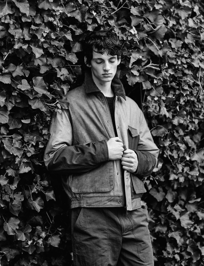 Monochrome portrait of a young adult male in a jacket posing in front of a lush ivy-covered wall in natural daylight.