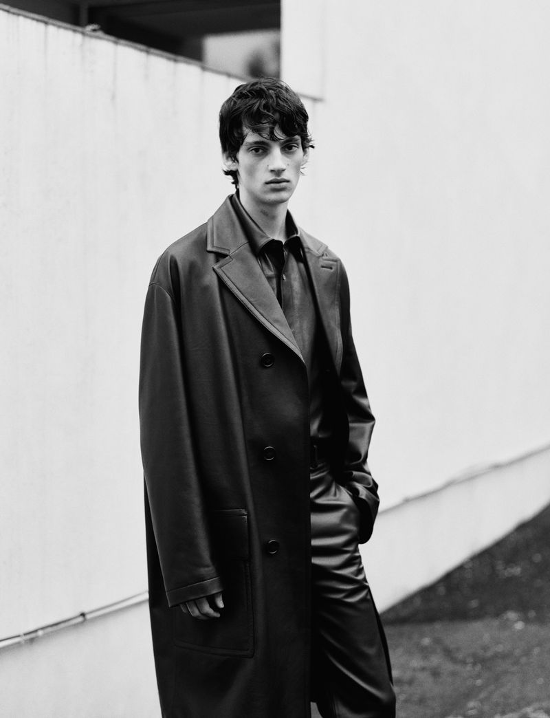 Young man with dark hair in a leather overcoat standing outside against a plain light wall in a monochrome portrait.