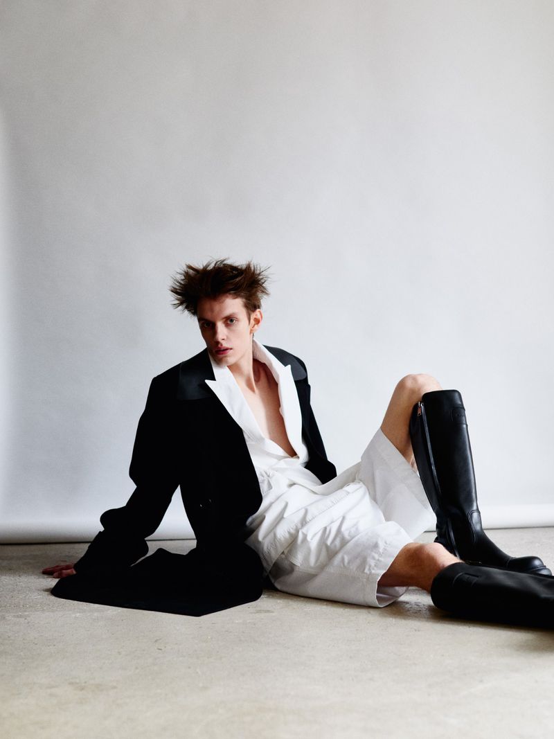 Young adult man in unbuttoned shirt and oversized blazer seated on the floor with knee-high leather boots in a fashion shot.