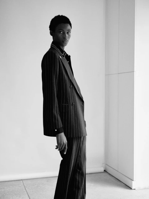 Monochrome portrait of a slender black man in a pinstripe suit, posing in front of a white wall, standing in a fashion photo studio.