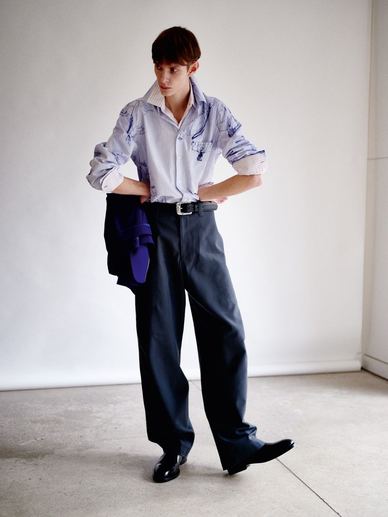 Fashionable young adult in collared shirt and wide-leg pants strikes a pose indoors with hands on hips against a neutral backdrop.