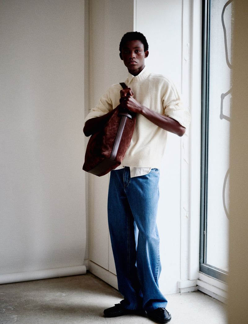 Dark-skinned male model in jeans, cream top, and a brown leather bag stands in front of a window in a neutral indoor setting.