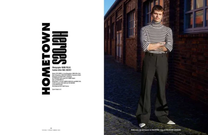A man wearing a striped turtleneck and black trousers standing in an alleyway with red brick buildings
