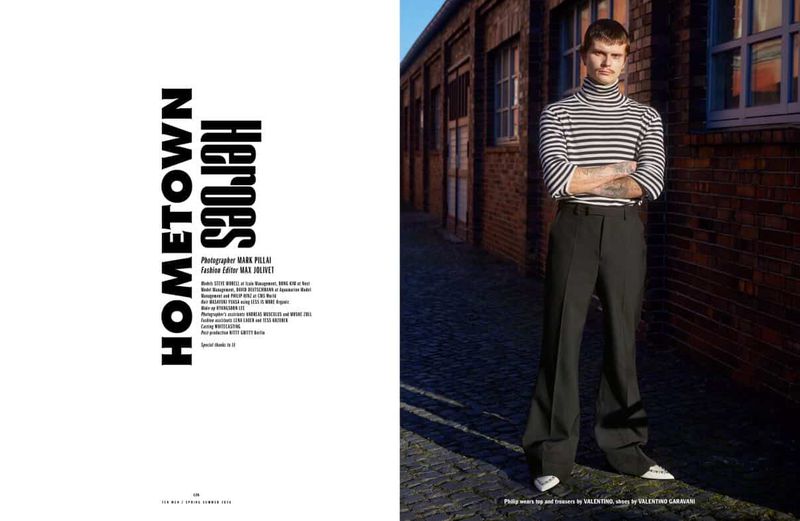 A male model poses in an urban setting, wearing a striped turtleneck and wide-leg black trousers, styled in a fashion editorial context.