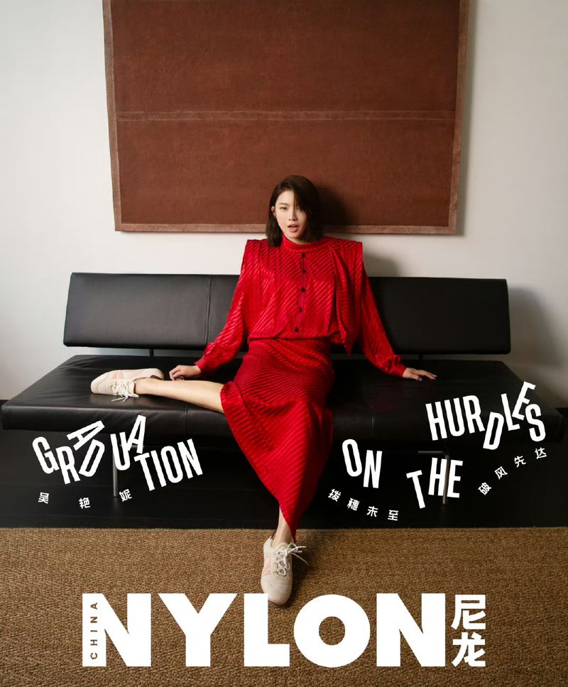 A stylish adult female poses on a black couch in a red dress for a fashion editorial with a painting and Nylon magazine branding.