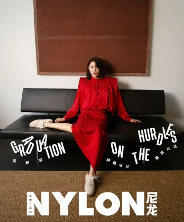 A stylish adult female poses on a black couch in a red dress for a fashion editorial with a painting and Nylon magazine branding.