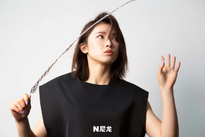 Portrait of a woman with short brown hair and a black dress, holding a patterned object, set against a clean white background.