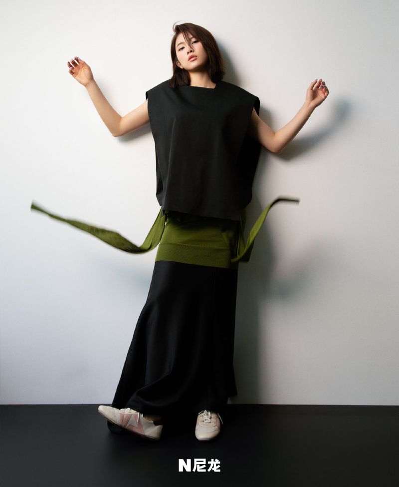 Fashionable woman in black sleeveless top, green wrap belt, and black skirt, posing against a white wall, showcasing modern, minimalist style.