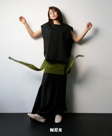 Fashionable woman in black sleeveless top, green wrap belt, and black skirt, posing against a white wall, showcasing modern, minimalist style.