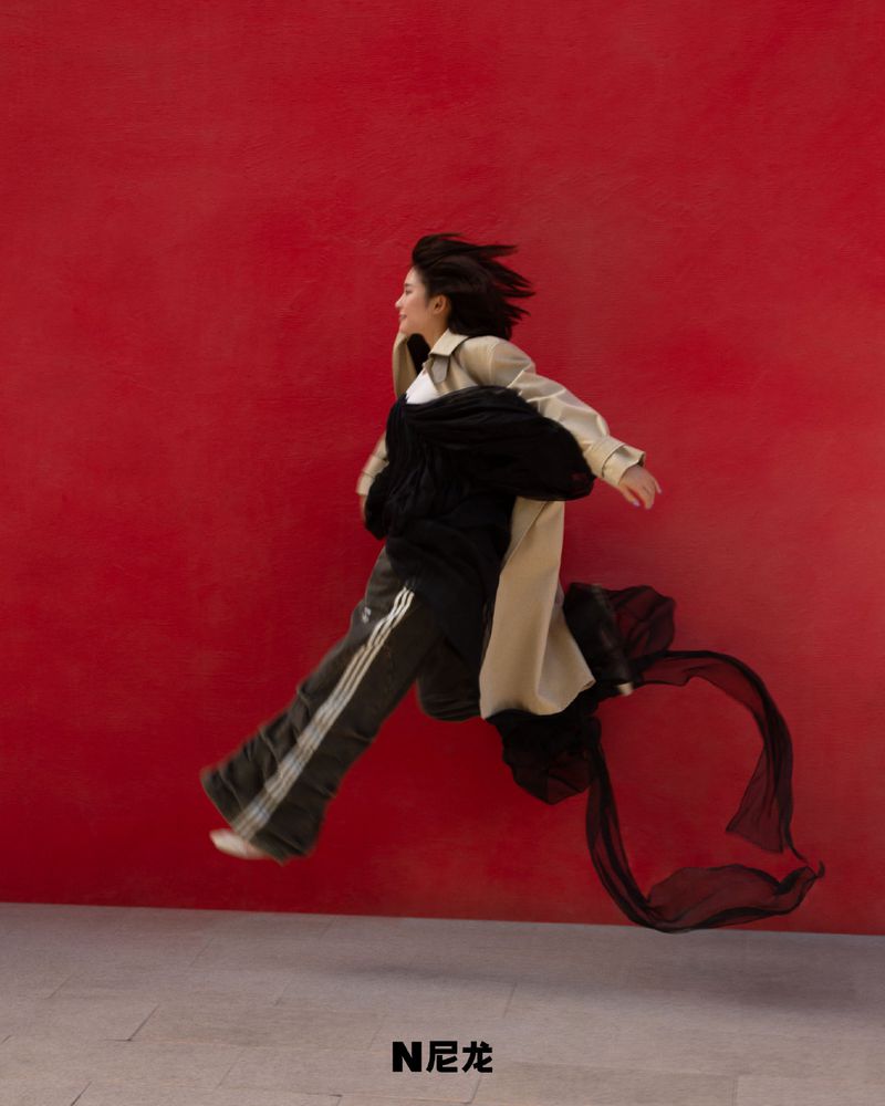Woman in motion, wearing a beige trench coat and black athletic pants with white stripes, set against a red wall and gray floor.