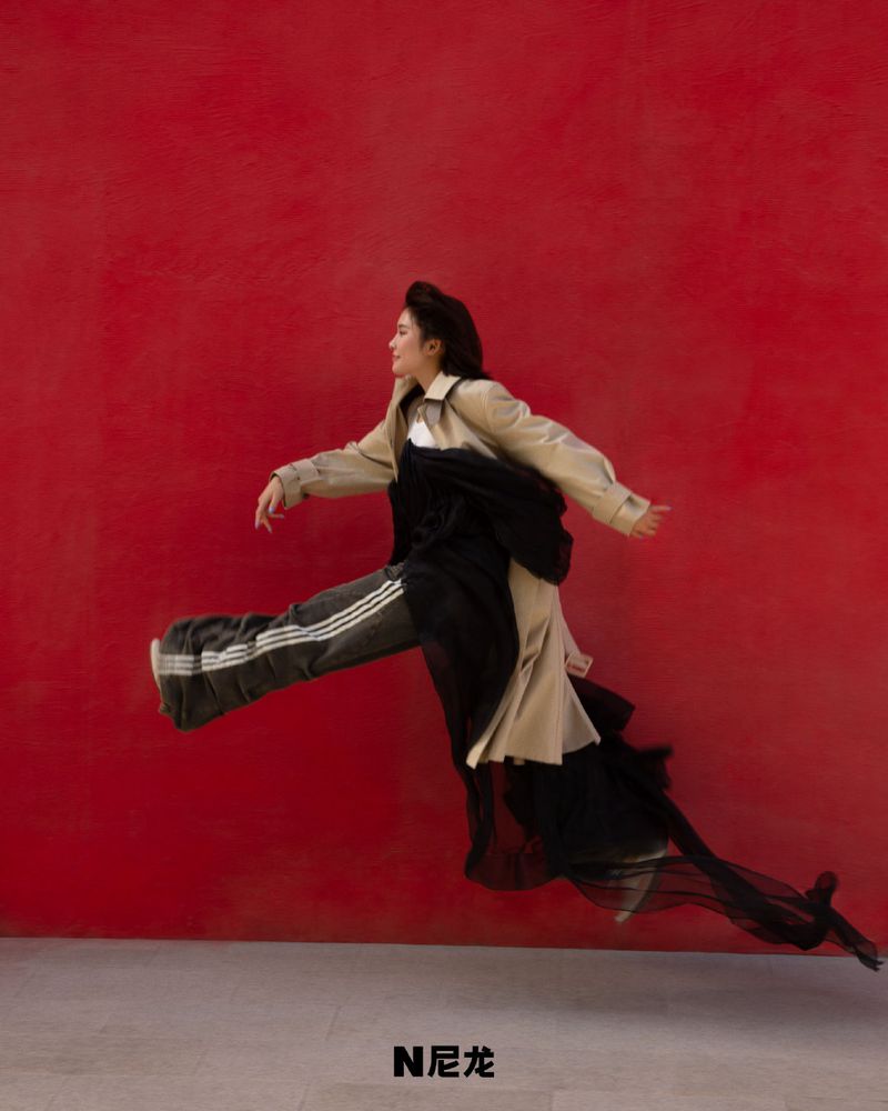 A young adult female in a beige trench coat and black pants with white stripes leaps in front of a bold red backdrop in a studio setting.