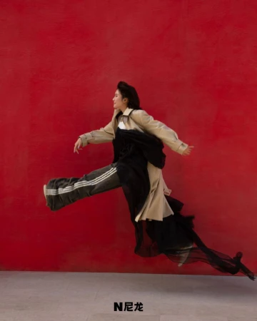 A young adult female in a beige trench coat and black pants with white stripes leaps in front of a bold red backdrop in a studio setting.
