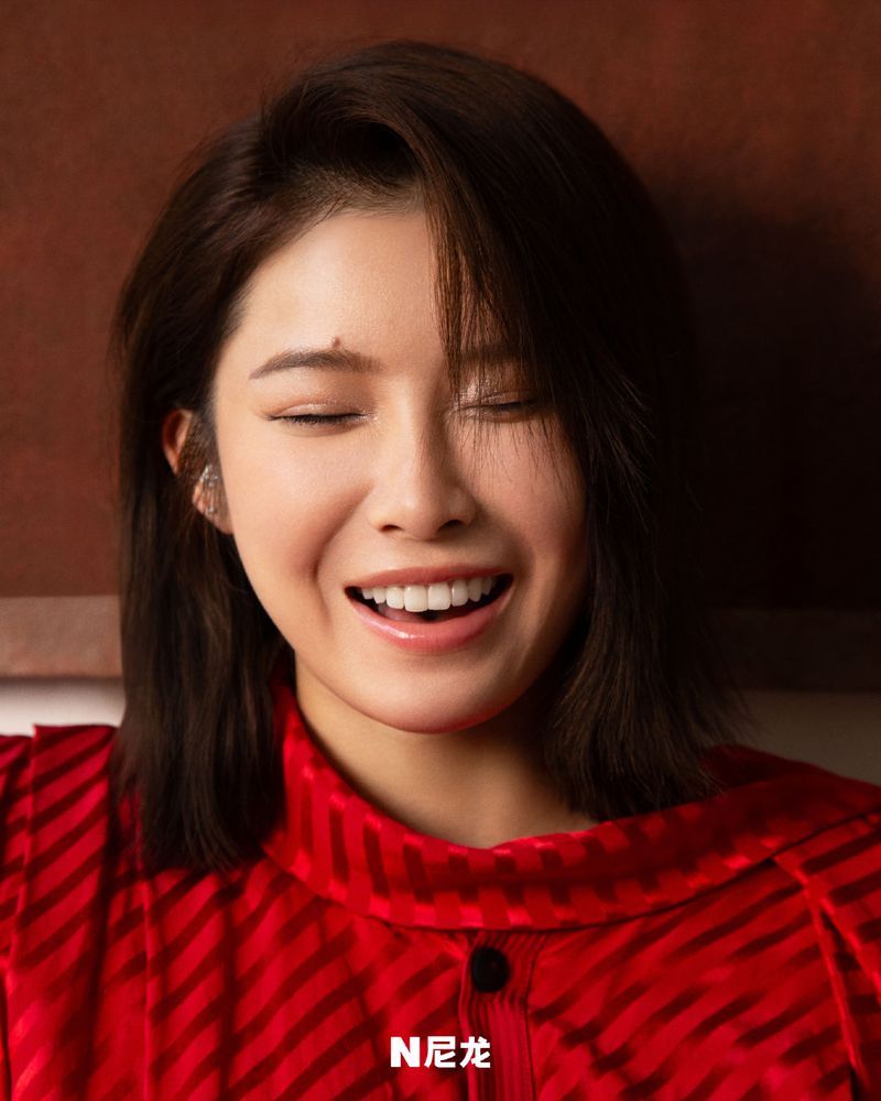 Close-up portrait of a smiling Asian woman with short dark hair, wearing a red patterned shirt, against a dark background.
