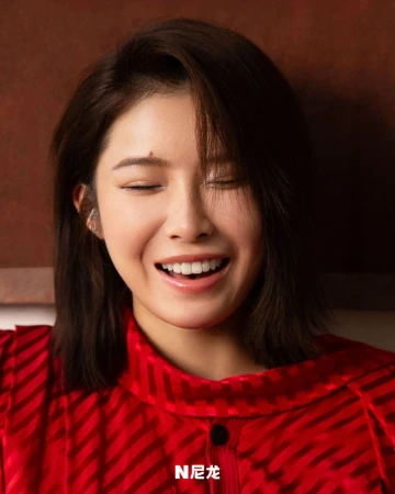 Close-up portrait of a smiling Asian woman with short dark hair, wearing a red patterned shirt, against a dark background.