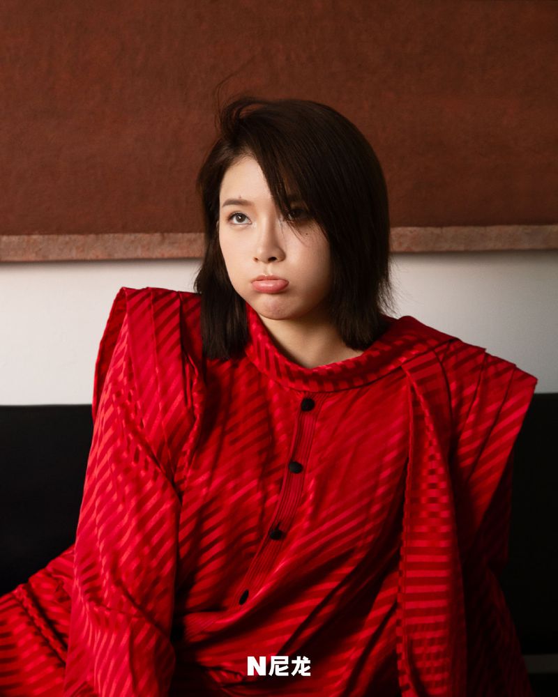 Portrait of an adult woman with dark hair and a pouting expression wearing a red textured dress sitting indoors with a simple background.