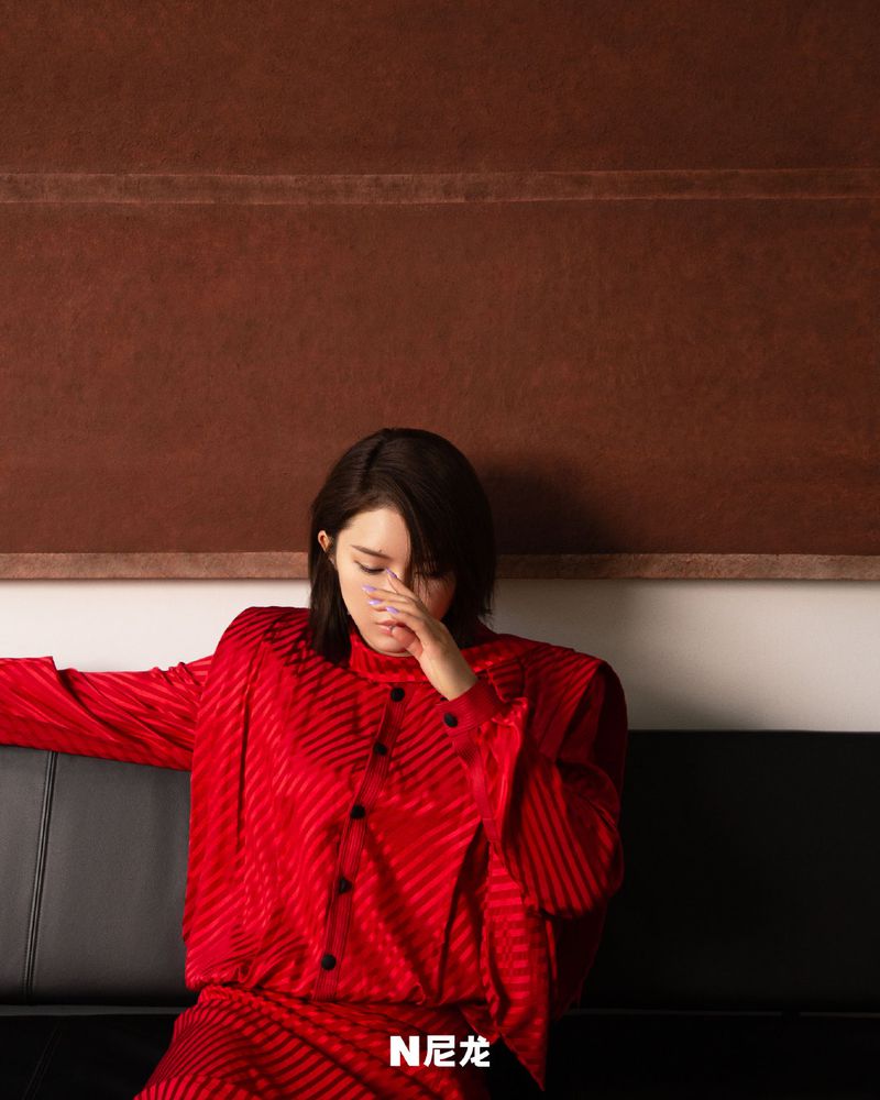 A woman with short dark hair sits on a couch wearing a red top and skirt in a studio setting with a brown backdrop.