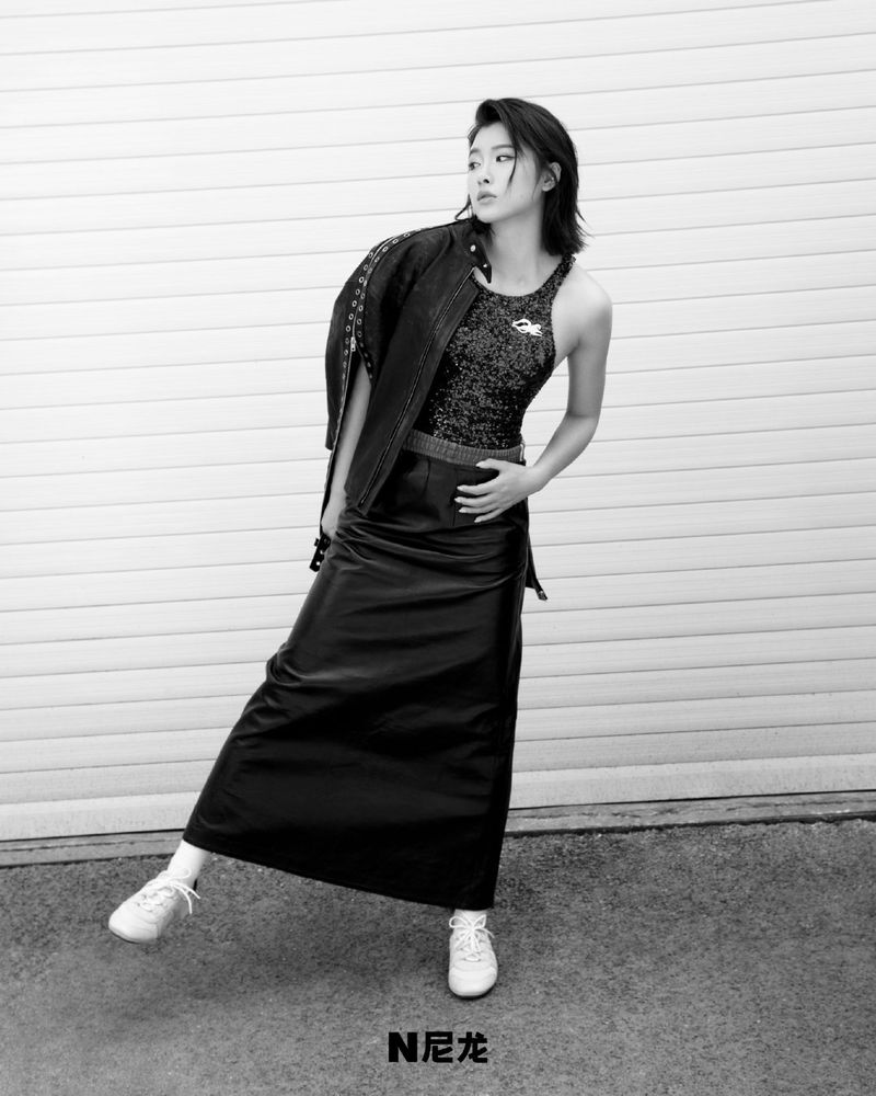 An Asian woman in a black leather skirt, tank top, and jacket strikes a casual pose against a textured white wall, showcasing fashion and style.