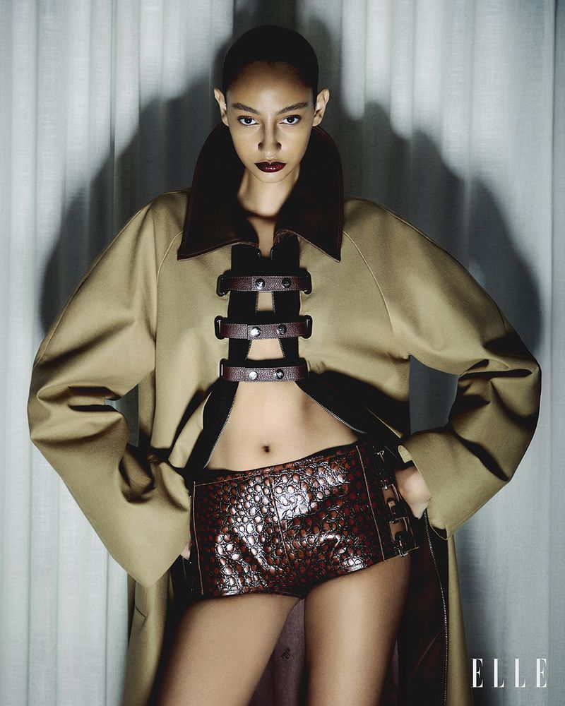 A striking fashion image of an adult female model in a beige trench coat and high-waisted dark brown textured shorts against a soft white backdrop.