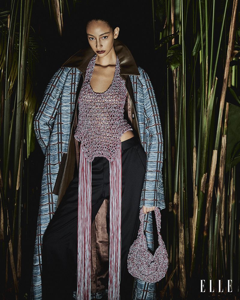 Fashion shot of a woman in a knit halter top and long coat, holding a matching knit bag, with tropical plants in the background.