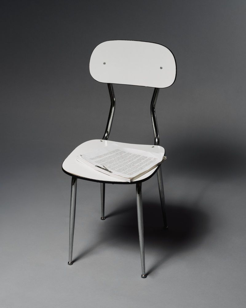 Minimalist photograph of a white chair with chrome legs and a document on the seat, set against a neutral gray backdrop.