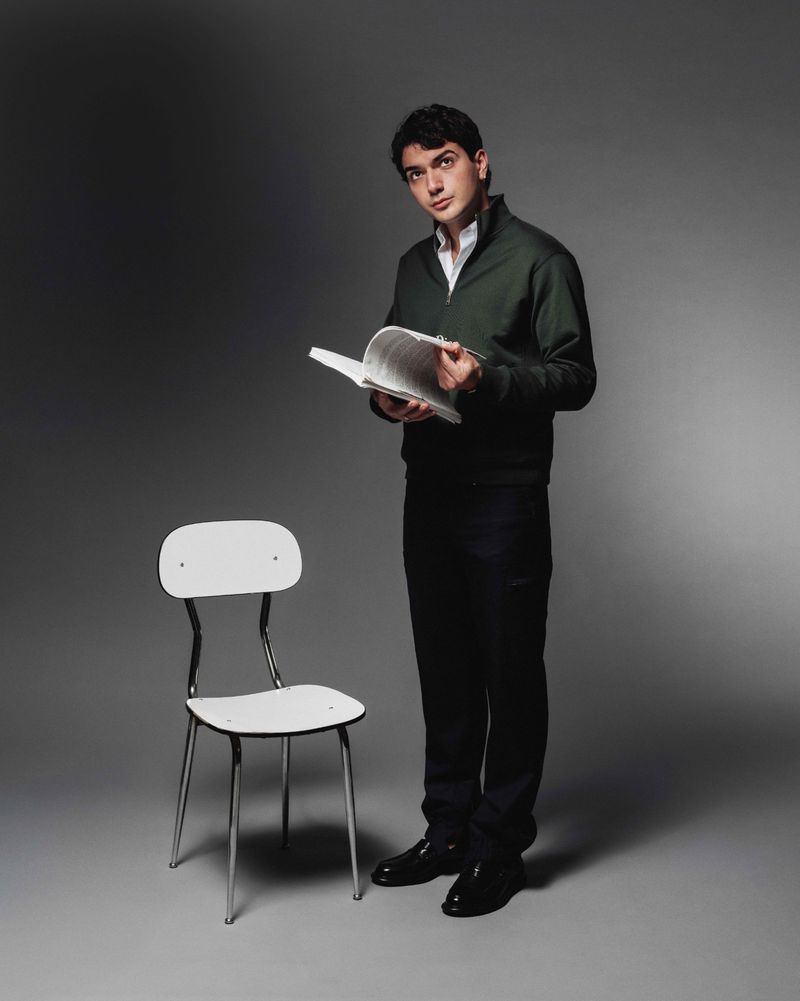 Studio shot of a young adult man reading a book while standing next to a white chair, with a grey background and medium framing.