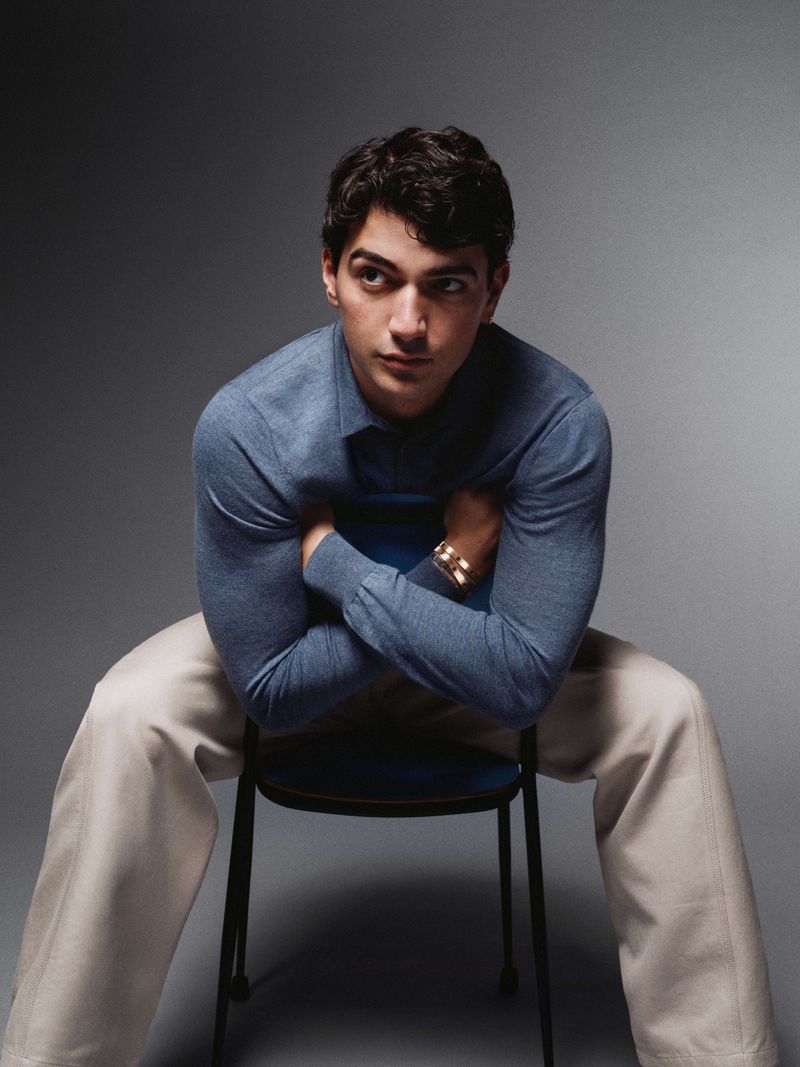 Portrait of a stylish adult man in a blue shirt and tan pants sitting on a blue chair in a photo studio with a grey background.