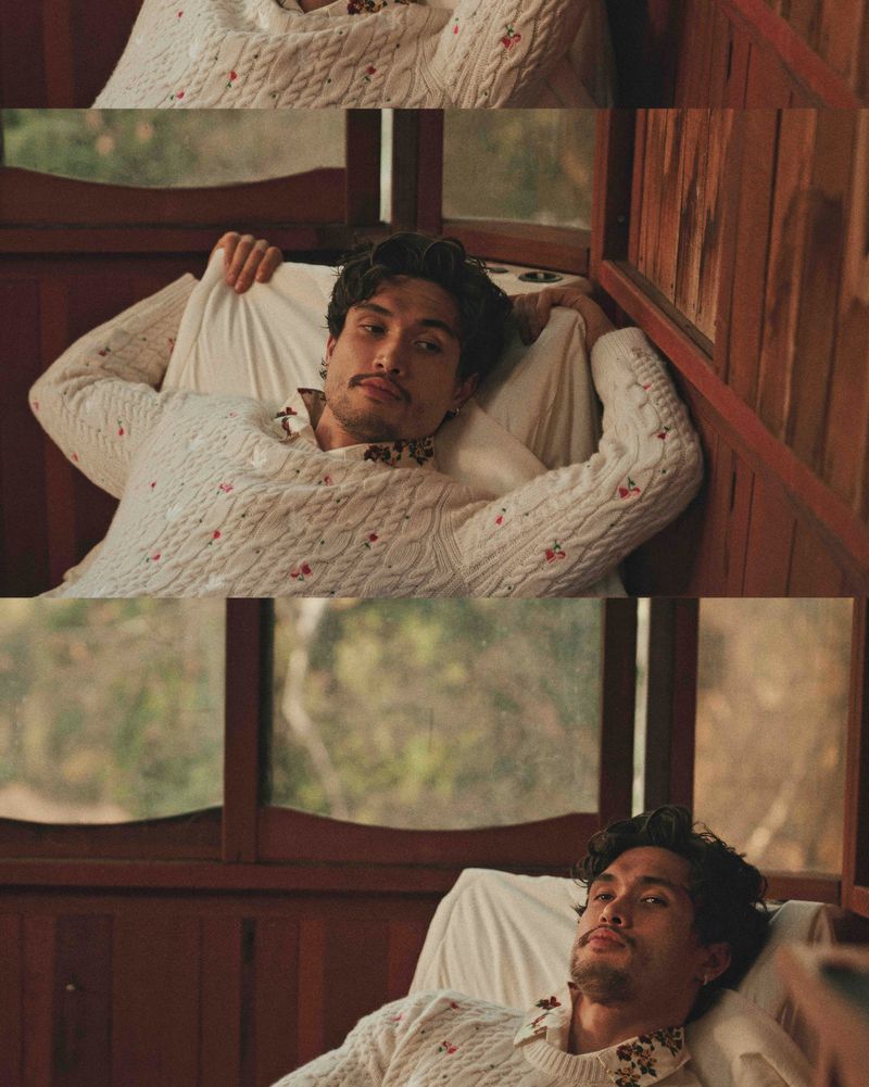 Adult male model poses indoors with white patterned sweater, window background. Casual, restful moment captured in soft, natural light.