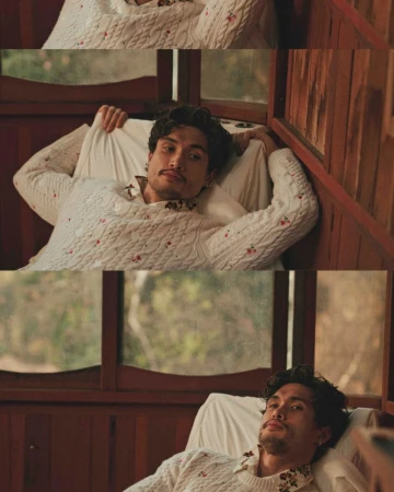 Adult male model poses indoors with white patterned sweater, window background. Casual, restful moment captured in soft, natural light.