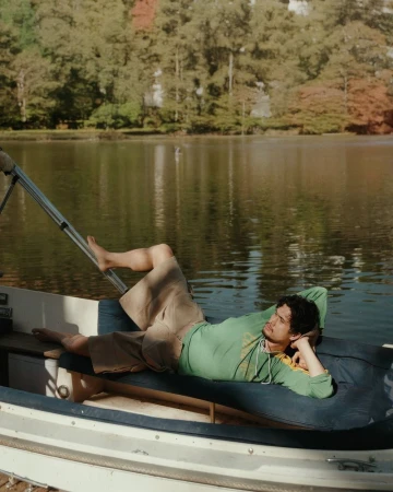 A man in a green shirt and tan shorts reclines on a blue cushion in a boat, with a calm lake and trees in the background.