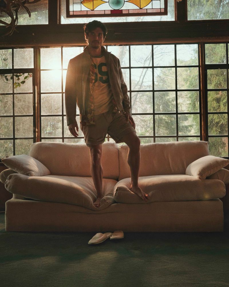 Man standing on a sofa in a sunlit room, wearing a jacket, t-shirt, and shorts, with bare feet, and a pair of shoes on the floor.