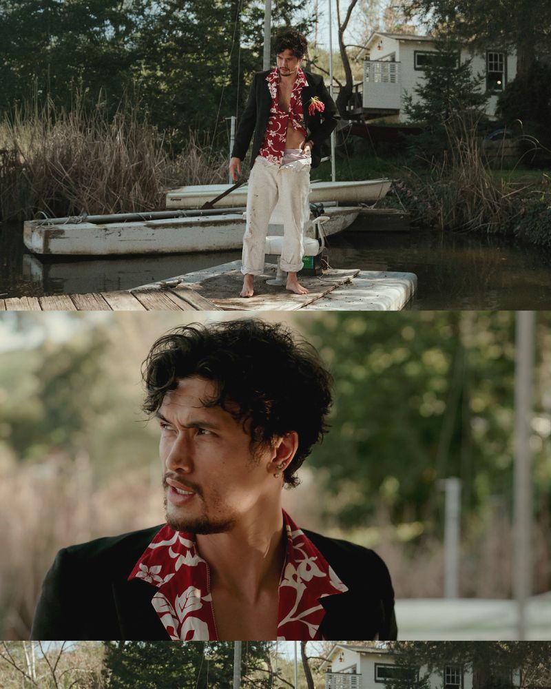 Man in black blazer and Hawaiian shirt stands on a dock near a lake, with boats and a white house visible in the background.