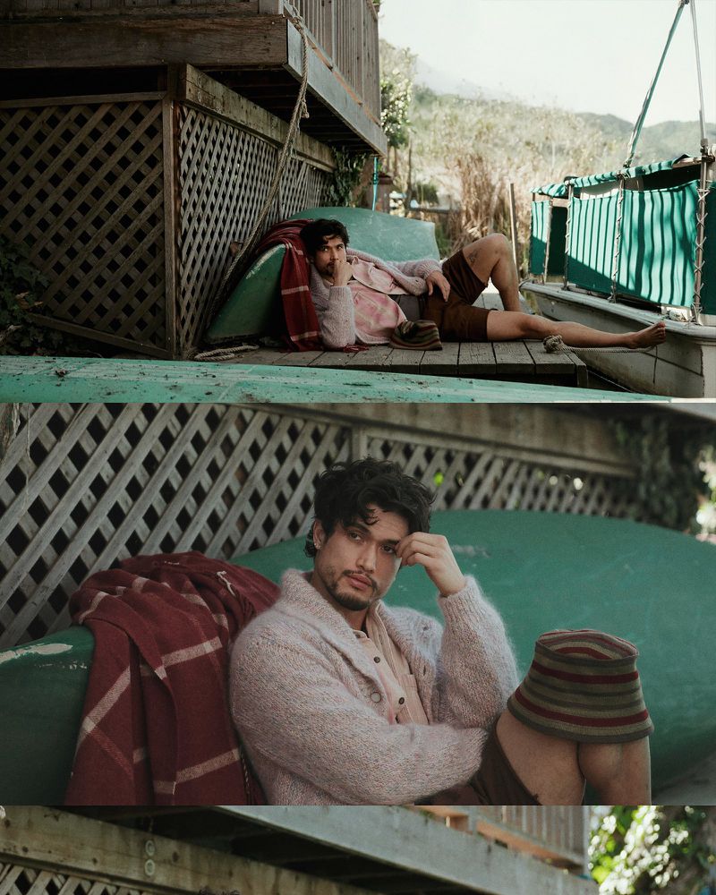 Adult male lies on a wooden porch in the daylight, resting on a green cushion with a patterned blanket, looking thoughtful in a cardigan.
