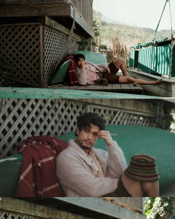 Adult male lies on a wooden porch in the daylight, resting on a green cushion with a patterned blanket, looking thoughtful in a cardigan.