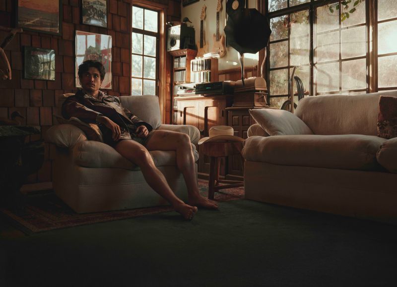 A man relaxes in an armchair in a sunlit room with vintage decor. The room has guitars on the wall and is warmly lit.