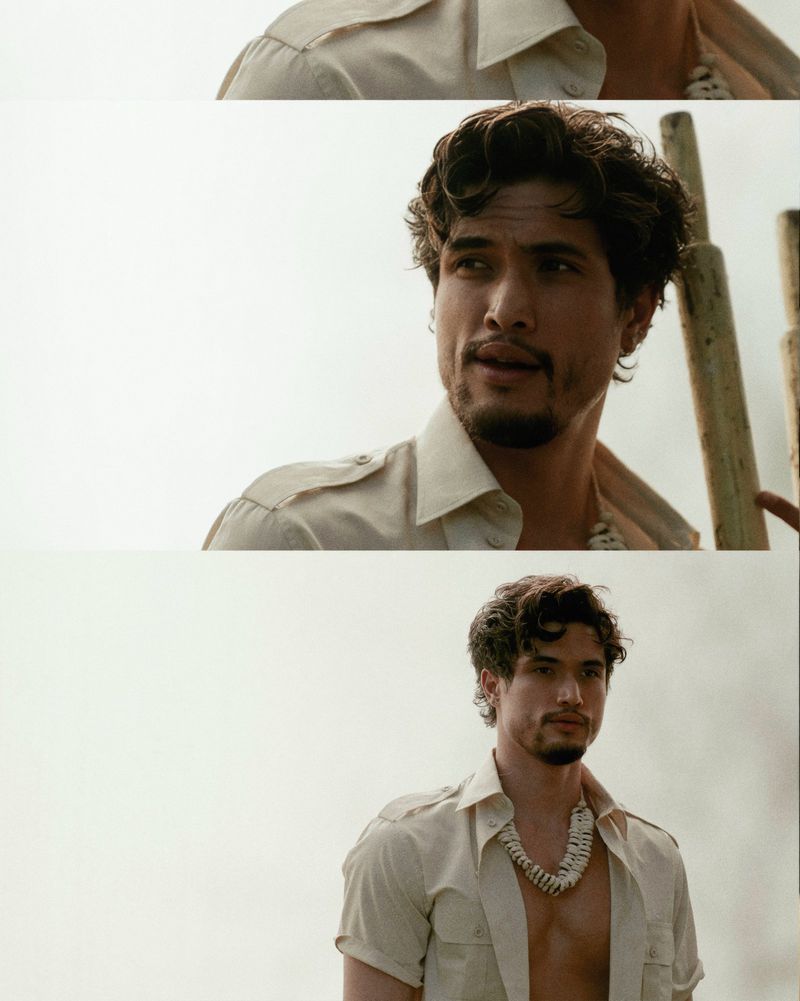 Portrait of a handsome man in a beige shirt with a shell necklace, looking off-camera in a bright outdoor setting.