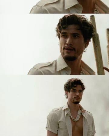 Portrait of a handsome man in a beige shirt with a shell necklace, looking off-camera in a bright outdoor setting.