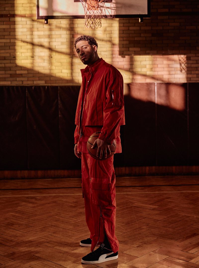 Man in red suit posing on basketball court holding basketball, lit by strong sunlight creating shadows on wall and floor.