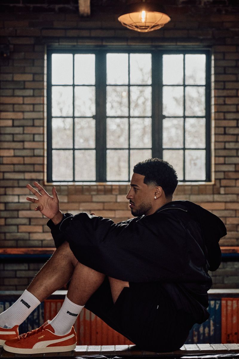 Adult male wearing a black hoodie and shorts seated with red sneakers in a brick-walled urban setting with a large window.