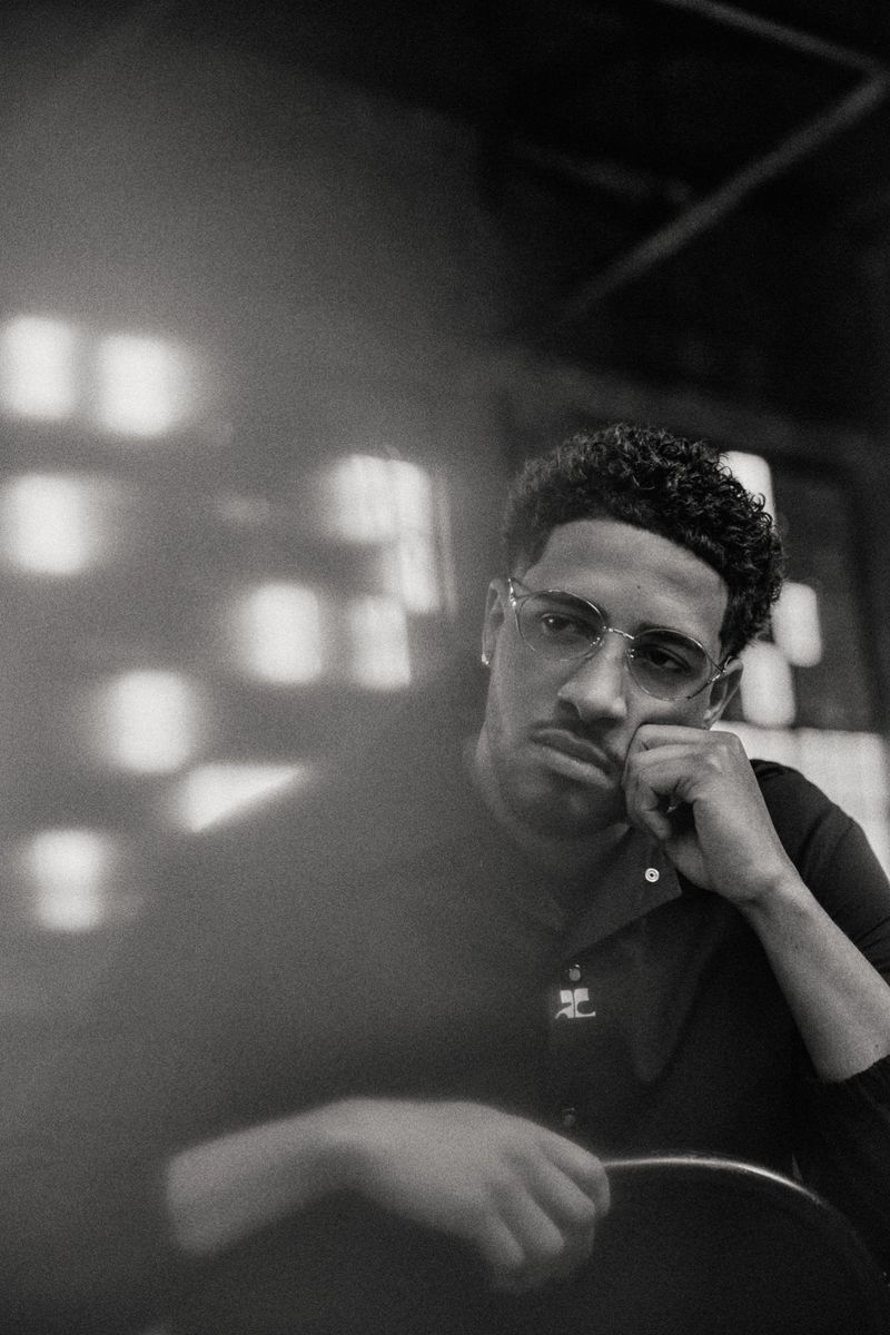 Monochrome portrait of adult male with glasses indoors. Man poses with curly hair, wearing black polo shirt, resting head on hand.